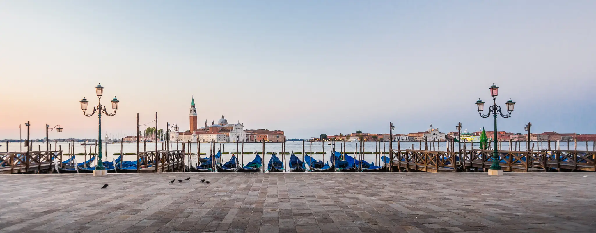 HOCHZEITSFOTOGRAF VENEDIG 33 Friedliche Morgenstimmung an einer venezianischen Uferpromenade mit aufgereihten blauen Gondeln, die auf das ruhige Wasser der Lagune und die Insel San Giorgio Maggiore bei Sonnenaufgang blicken.