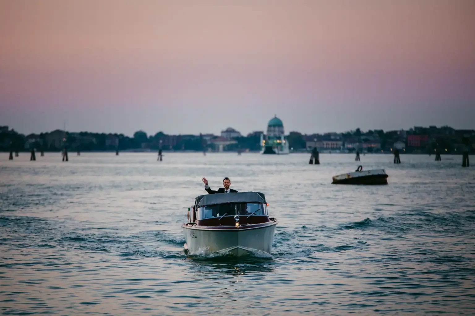 Ein elegant gekleideter Bräutigam winkt freudig aus einem klassischen venezianischen Wassertaxi aus Mahagoniholz, das während einer exklusiven Hochzeitsreise über die weite Lagune von Venedig fährt.