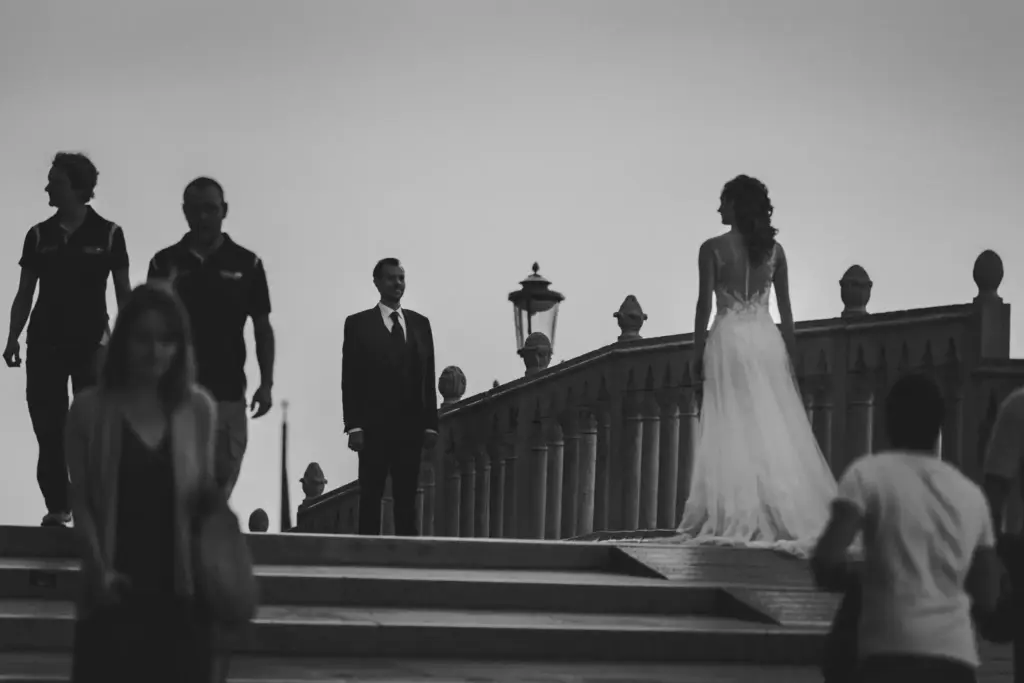 Filmische Schwarz-Weiß-Szene im Reportagestil: Braut und Bräutigam gehen als elegante Silhouetten über eine venezianische Brücke, umgeben vom erwachenden Leben der Stadt.