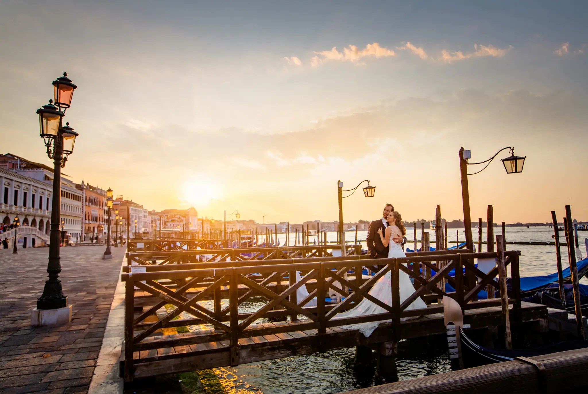 HOCHZEITSFOTOGRAF VENEDIG 44 Romantischer Moment eines Brautpaares auf einem hölzernen Bootssteg in Venedig, sanft beleuchtet vom frühen Sonnenlicht, mit typischen blauen Gondeln im Hintergrund.
