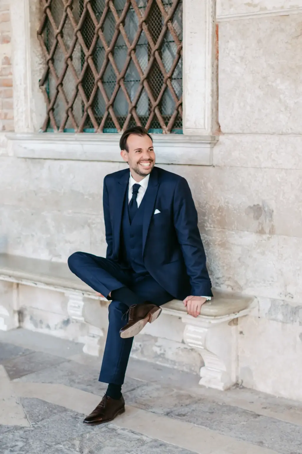 Lässiges und elegantes Porträt des Bräutigams im blauen Maßanzug, der entspannt an einer historischen Steinwand in Venedig sitzt.