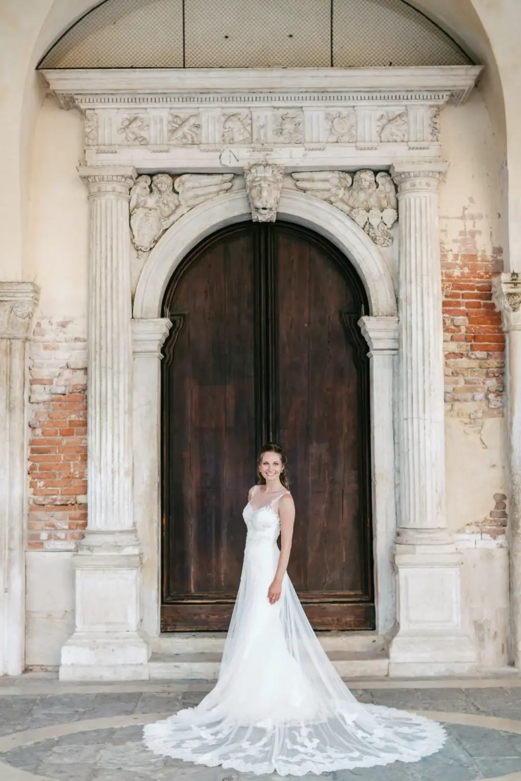 Selbstbewusstes und natürliches Porträt einer lachenden Braut in einem fließenden Hochzeitskleid, fotografiert vor einem massiven historischen Holzportal mit antiken Steinmetzarbeiten in Venedig.