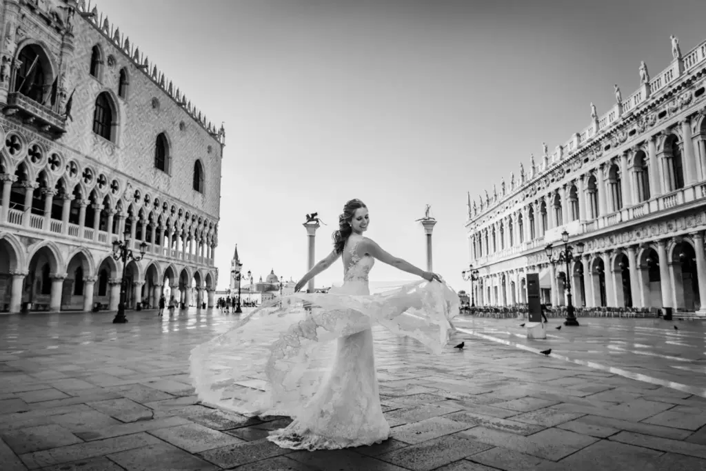 Schwarz-Weiß-Aufnahme einer Braut, die ihr Brautkleid auf dem leeren Markusplatz in Venedig schwingen lässt, flankiert vom Dogenpalast und der Markusbibliothek im frühen Morgenlicht.