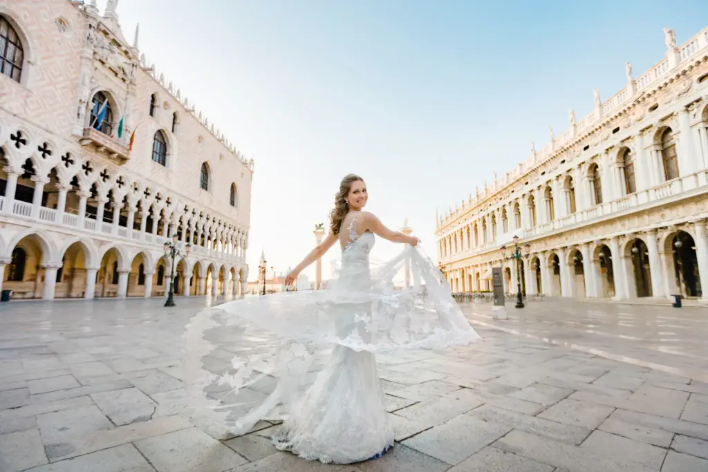 Glückliche Braut präsentiert ihr fließendes Spitzenkleid auf dem morgendlich leeren Markusplatz vor der Kulisse des Dogenpalastes. Ein klassisches Motiv beim After Wedding Shooting in Venedig.