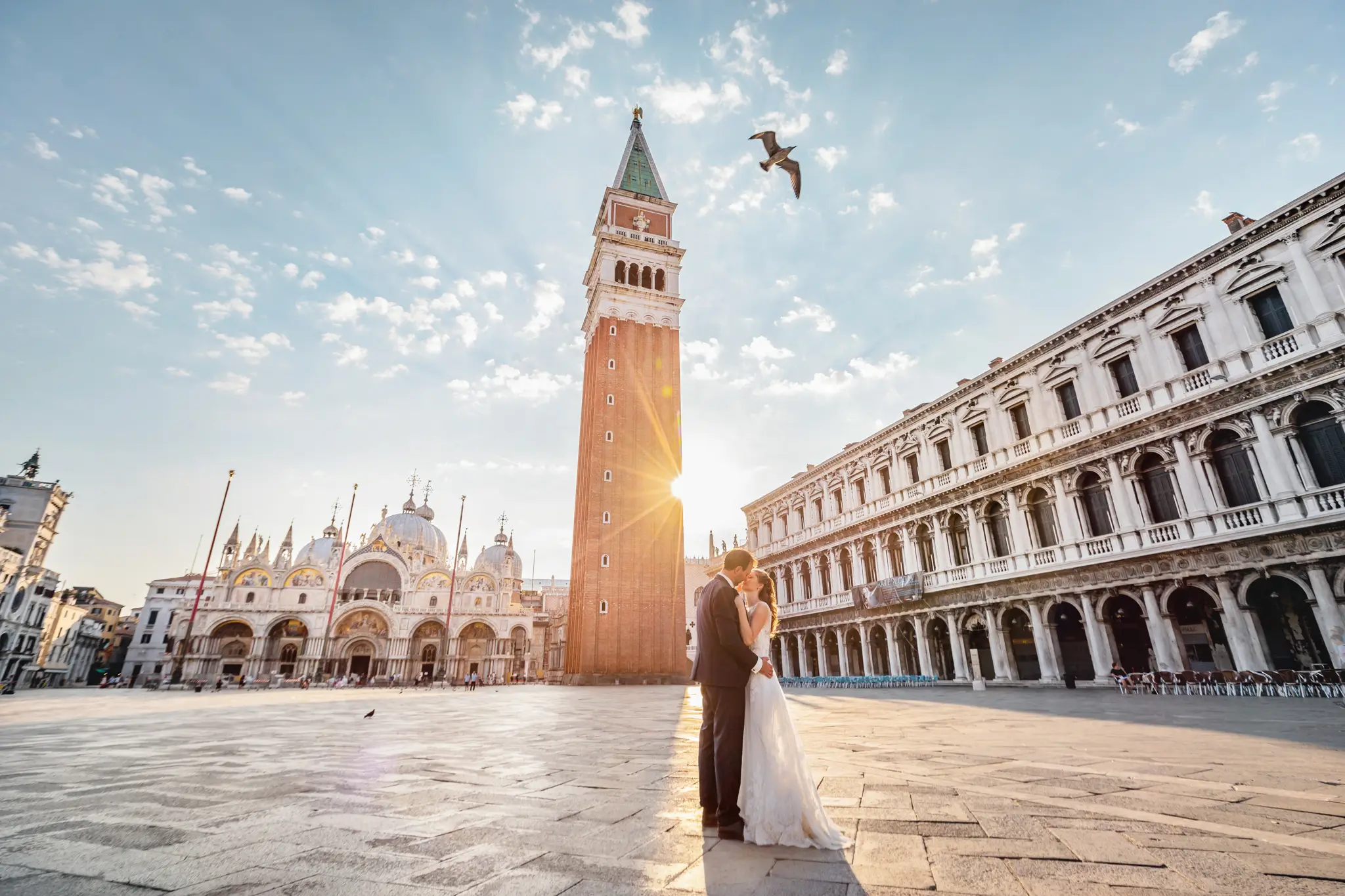 HOCHZEITSFOTOGRAF VENEDIG 53 Brautpaar küsst sich vor dem Campanile di San Marco, während die aufgehende Sonne als Sonnenstern hinter dem Glockenturm hervorbricht. Hochzeitsfotos im goldenen Morgenlicht von Venedig.