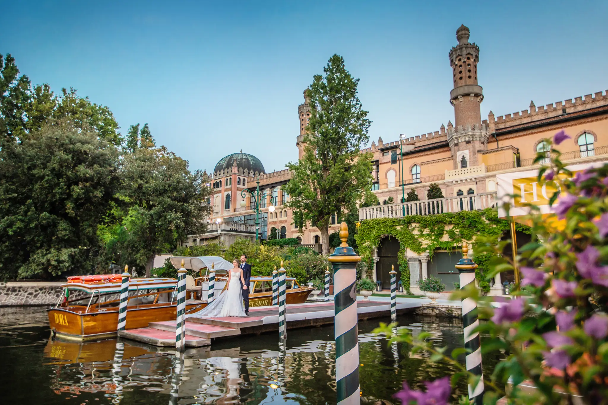 HOCHZEITSFOTOGRAF VENEDIG 58 Brautpaar an Hand am Bootssteg vor dem orientalisch anmutenden Hotel Excelsior Venice Lido, im Wasser ein klassisches venezianisches Holzboot. Hochzeitsfotos abseits der Touristenpfade.