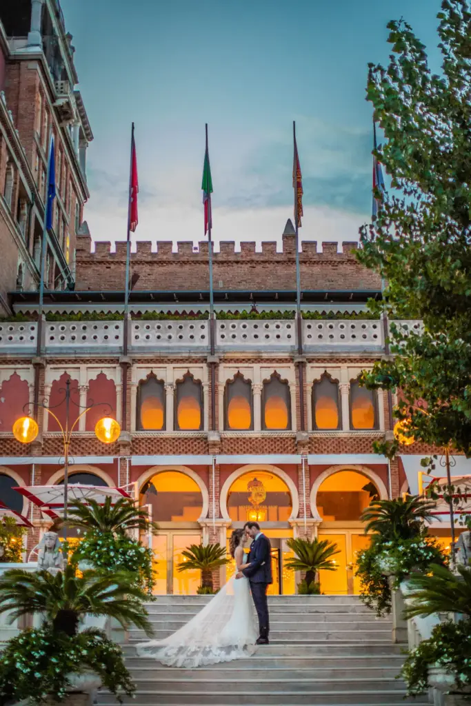 Brautpaar küsst sich auf der prachtvollen Eingangstreppe des Hotel Excelsior Venice Lido im warmen Abendlicht der Fackeln. Destination Wedding in Venedig zur blauen Stunde.
