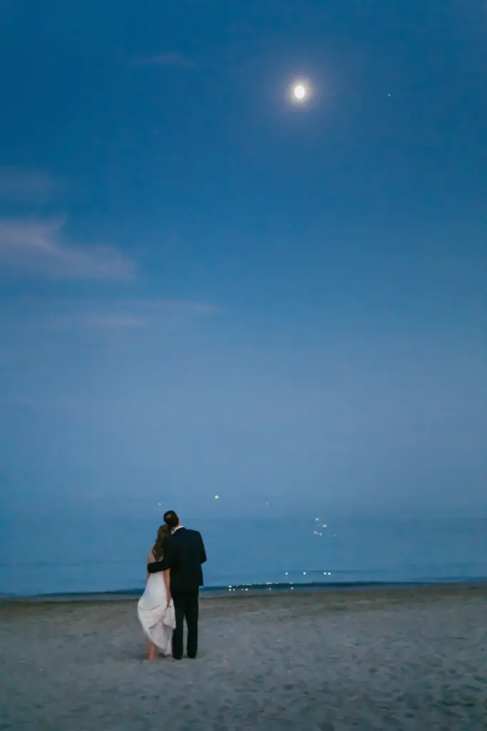 Brautpaar steht eng umschlungen am nächtlichen Strand des Lido di Venezia und blickt auf das mondbeschienene Meer, der letzte stille Moment des Tages.